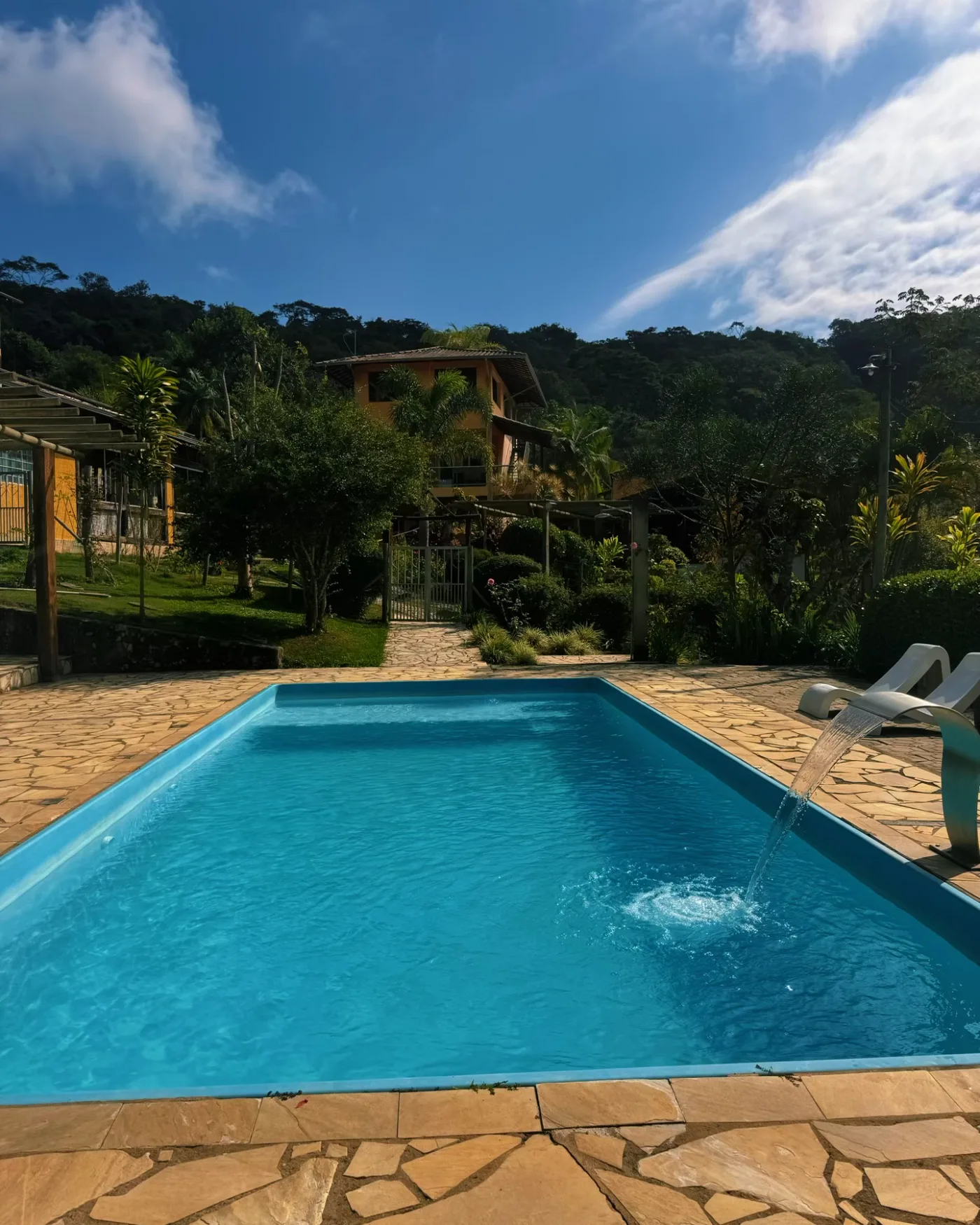 Piscina do sítio com cascata ligada, deck de pedra e a mata ao fundo